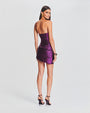 A backside full body view of a brunette model with her hair swept to the side wearing a strapless, shiny purple dress with ruched detailing all across it and an open-back design. The model is also wearing purple high-heels and gold earrings, while standing in front of a white background.