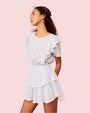 An angled view of a curly-haired brunette model wearing a white dress with ruffled sleeves, a tiered skirt, and a fitted bodice with intricate ribbed detailing. The background is pink