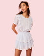 A curly-haired model with her hands behind her back, wearing a white dress with ruffled sleeves, a tiered skirt and a fitted bodice with intricate ribbed detailing. The background is pink.
