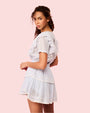An angled backside view of a curly-haired model wearing a white dress with ruffled sleeves, a tiered skirt and a fitted bodice. The background is pink.