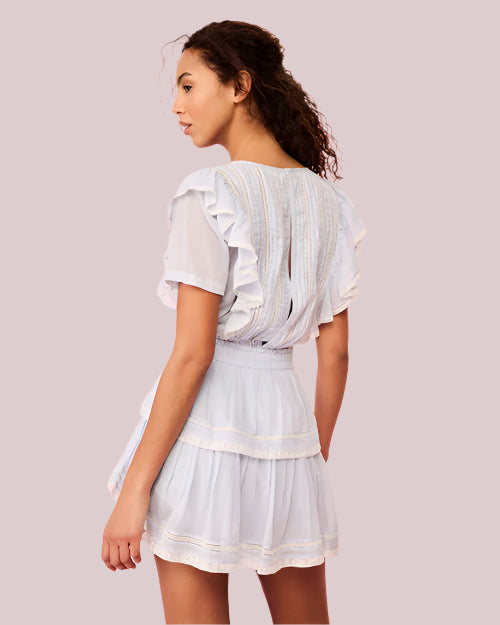 A backside view of a curly-haired model wearing a white dress with ruffled sleeves, a tiered skirt and a fitted bodice with a ribbed design. The background is pink.