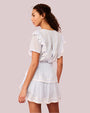 A backside view of a curly-haired model wearing a white dress with ruffled sleeves, a tiered skirt and a fitted bodice with a ribbed design. The background is pink.