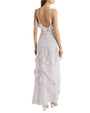 The backside view of a model wearing a long, sleeveless dress with thin straps, a subtle blue floral pattern and ruffles along the across the front leg area, the hem, and at the low neckline, making the dress backless. The model is wearing white heels in front of a white background.