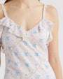 A close-up chest level image of a model wearing a sleeveless dress with thin straps, a subtle blue floral pattern and features ruffles along the neckline, and lace detailing along the body.