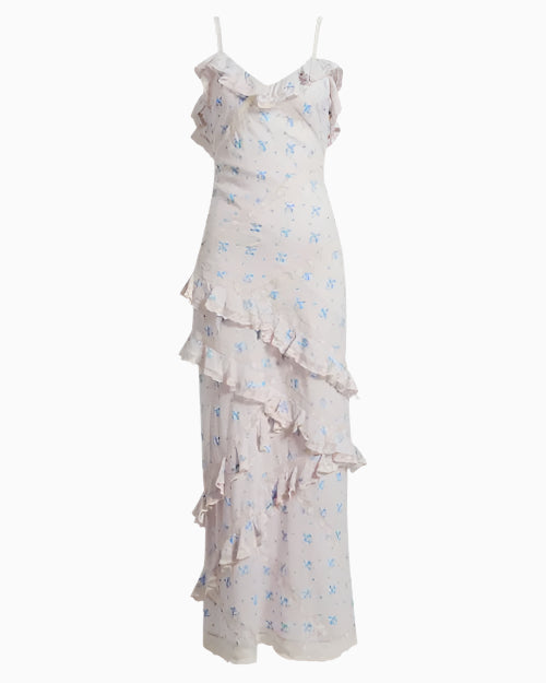 A full image of a long, sleeveless dress with thin straps. The dress is white with a subtle blue floral pattern and features ruffles along the neckline, across the front leg area, and at the hem.