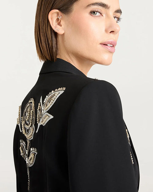 Close up of a back view of a person wearing a black jacket with intricate metallic floral embroidery, including roses, accented with beads or sequins.