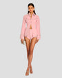 A complete frontal view of a brunette model wearing a coordinating set of a long-sleeved button down pink top and shorts. The top is buttoned at the center, with silver studs outlining the collar, creating a horizontal line across the upper chest, hemline, and bordering the cuffs. The model is also wearing large silver earrings and silver open-toed heels while standing in front of a white background.