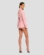 The backside view of a model wearing a coordinating set of a long-sleeved button down pink top and shorts. The top has silver studs outlining the collar, creating a horizontal line across the upper back, hemline and bordering the cuffs. The model is also wearing large silver earrings and silver open-toed heels, while standing in front of a white background.