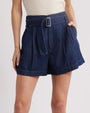 A close waist-level shot of a model with her hand in her pocket, wearing a white shirt tucked into high-waisted, dark blue denim shorts with pleats, and an attached denim belt featuring a vertical rectangular buckle.