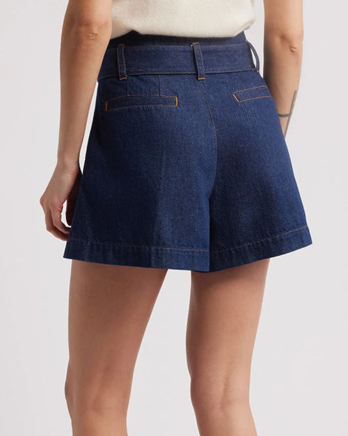 A close waist-level view of a model wearing a white shirt tucked into a pair of high-waisted, dark blue denim shorts with an attached denim belt. The background is white.