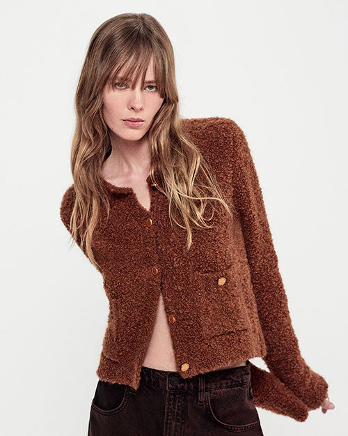 A waist-up view of a blonde model with her arms stretched behind her back wearing a brown, fuzzy textured cardigan featuring a high round neckline, gold buttons, and two waist pockets, as well as brown jeans. The model is standing against a white background.