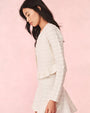 A side view of a dark-haired model wearing an open, white, long-sleeved cardigan over a matching white dress. The cardigan has a horizontal lace striped texture and frills at the waist. The background is plain pink.