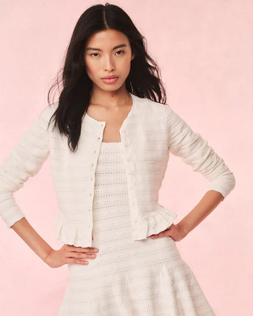 A torso shot of a dark-haired model with her hands on her hips, wearing an open, white, long-sleeved cardigan over a matching white dress. The cardigan has a horizontal lace striped texture and frills at the waist. The background is plain pink.
