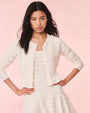 A torso shot of a dark-haired model with her hands on her hips, wearing an open, white, long-sleeved cardigan over a matching white dress. The cardigan has a horizontal lace striped texture and frills at the waist. The background is plain pink.