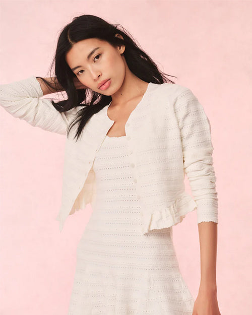 A torso shot of a dark-haired model with one hand on her head, wearing an open, white, long-sleeved cardigan over a matching white dress. The cardigan has a horizontal lace striped texture and frills at the waist. The background is plain pink.