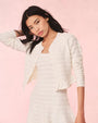 A torso shot of a dark-haired model with one hand on her head, wearing an open, white, long-sleeved cardigan over a matching white dress. The cardigan has a horizontal lace striped texture and frills at the waist. The background is plain pink.