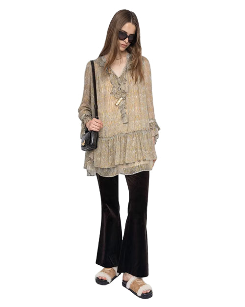 Person standing and wearing a loose, long-sleeved, floral-patterned blouse with ruffles and a layered hem, paired with black flared pants and white sandals with black straps. A black shoulder bag is worn.