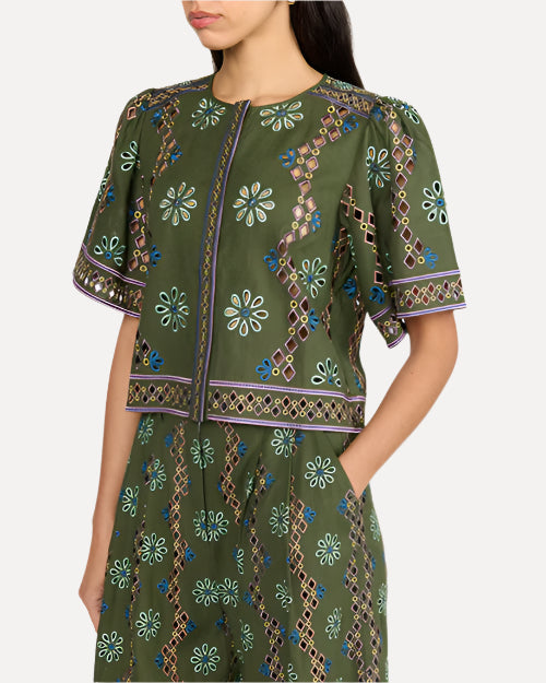 A close-up body shot of a dark-haired model with her hand in her pocket, standing against a white background and wearing a matching green short-sleeved top and bottoms with intricate patterns, adorned with floral and geometric designs.