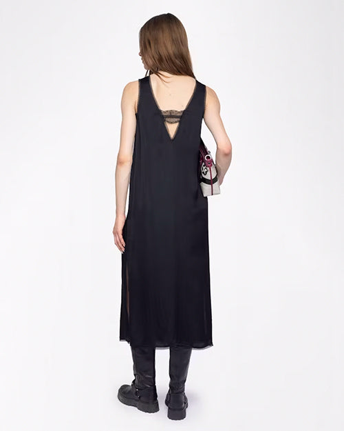 A backside view of a brunette model wearing a long, sleeveless black dress with a deep V-neckline and lace detailing across the back opening. She is holding a colorful handbag and chunky black boots, while standing against a white background.