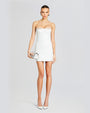 A full body view of a brunette model with her hair tied back, wearing a strapless white mini dress with a straight neckline and a straight lacey neckline. The model is also wearing a silver clutch and white pointed-toe heels. The background is white.
