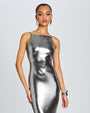 A knee-up full body view of a brunette model wearing a floor-length, metallic silver dress with
thin straps. The model is wearing silver earrings, a silver bracelet and is standing in front of a white background.