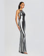An angled full body view of a brunette model wearing a floor-length, metallic silver dress with a backless design and thin straps. The model is wearing silver earrings, open-toed heels and is standing in front of a white background.