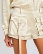 A close-up image of a models' hip wearing a beige satin shirt tucked into matching shorts, with utility style pockets and flaps on both front sides and long strings. The background is white.