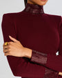 A zoomed in chest-level view of a model with her hand holding her arm wearing a gold ring and a dark red dress with long sleeves, a high neckline, and a shiny texture on the skirt, cuffs, and neck.