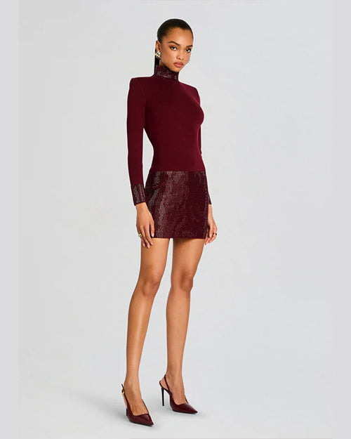 An angled full body view of a model wearing a dark red dress with long sleeves, a high neckline, and a shiny texture on the skirt, cuffs, and neck. The model is also wearing matching dark red high-heeled shoes and standing in front of a blank background.