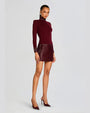 An angled full body view of a model wearing a dark red dress with long sleeves, a high neckline, and a shiny texture on the skirt, cuffs, and neck. The model is also wearing matching dark red high-heeled shoes and standing in front of a blank background.