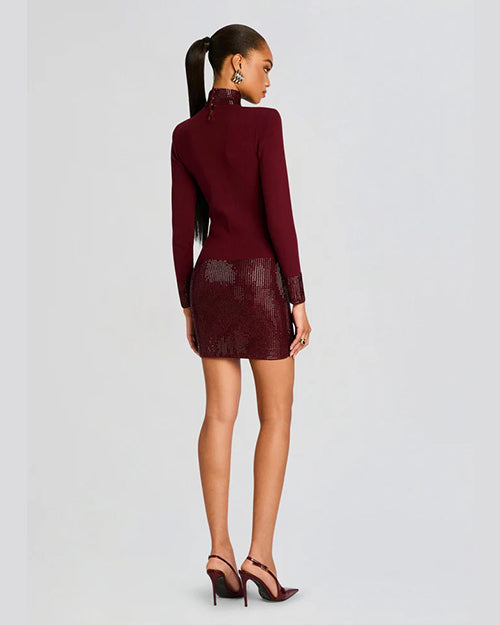 A backside full body view of a model with a high ponytail wearing a dark red dress with long sleeves, a high neckline, and a shiny texture on the skirt, cuffs, and neck. The model is also wearing matching dark red high-heeled shoes and standing in front of a blank background.