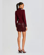 A backside full body view of a model with a high ponytail wearing a dark red dress with long sleeves, a high neckline, and a shiny texture on the skirt, cuffs, and neck. The model is also wearing matching dark red high-heeled shoes and standing in front of a blank background.