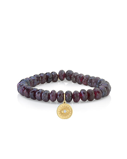 A wine-red beaded bracelet with a gold circular charm featuring an eye with a jewel in the middle. The background is white.