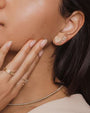 A close-up of a brunette models' jaw with her hand on her cheek, wearing two earrings—one shaped like a heart adorned with jewels and the other designed as an arrow and feather. The model is also wearing two gold rings and a silver chain necklace.