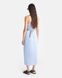 The backside view of a brunette model wearing a light blue shin length dress with thin straps. The dress has light blue fabric tied around the waist. The person is also wearing black sandals and sunglasses to match.