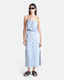 A brunette model with black sunglasses wearing a long light blue dress with thin straps. The dress has buttons running down the front and light blue fabric tied and knotted at the waist. The person is also wearing black sandals and standing against a white background.