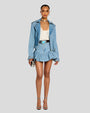 A full-frontal view of a brunette model wearing a light blue denim jacket with a large collar, long sleeves, a zipper closure, and a belt at the waist. The jacket is unzipped, and paired with a matching short, pleated skirt, a white low-neck shirt, a leather belt and silver earrings, and high heels.