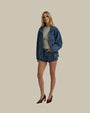 A complete frontal view of a blonde model with her hand in her pocket, wearing blue denim shorts and a gray elbow-length top with a blue denim jacket and brown heels. The background is plain.