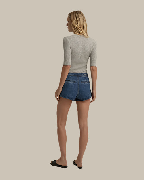 A full body backside view of a blonde model wearing blue denim shorts with "Favorite Daughter" embroidered on the waistband on the back. The person is also wearing a gray elbow-length top with black slides, standing against a grey background.