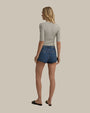 A full body backside view of a blonde model wearing blue denim shorts with "Favorite Daughter" embroidered on the waistband on the back. The person is also wearing a gray elbow-length top with black slides, standing against a grey background.