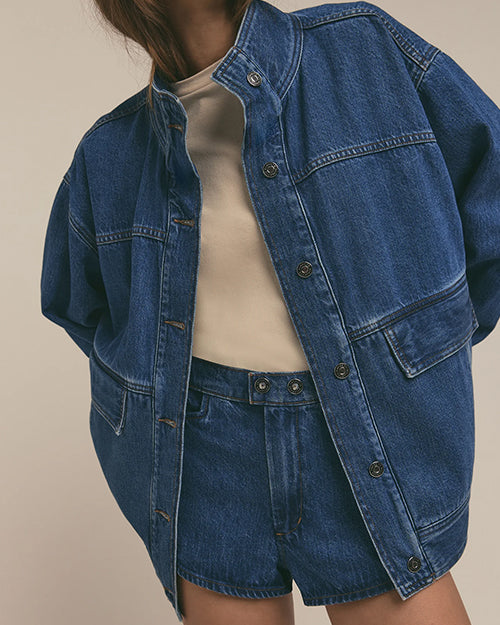 A close-up view of a model wearing a denim jacket with horizontal seams and a popped collar, with denim shorts and a tucked in cream shirt. The background is plain.