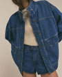 A close-up view of a model wearing a denim jacket with horizontal seams and a popped collar, with denim shorts and a tucked in cream shirt. The background is plain.