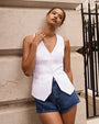 A model with her hand up to her neck, leaning against a stone wall, and wearing a white sleeveless vest with buttons down the front and blue high-waisted denim shorts.