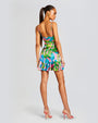 A full body backside view of a brunette model wearing a vibrant tropical print mini dress with spaghetti straps and an open back. She is also wearing a pair of gold earrings and pink high heels. The background is plain white.