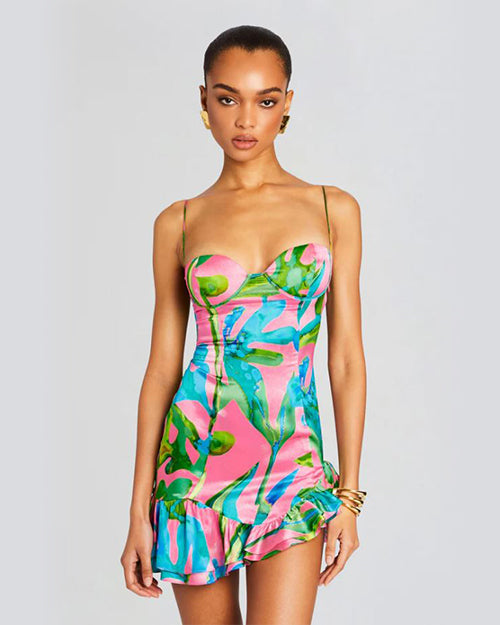 A knee-up view of a brunette model wearing a vibrant tropical print mini dress with spaghetti straps and a sweetheart neckline. She is wearing three gold bangles and large gold earrings. The background is plain white.