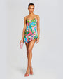A full body shot of a brunette model wearing a vibrant tropical print mini dress with spaghetti straps and a sweetheart neckline. She is holding a pink clutch and wearing pink heels with gold bracelets and earrings. The background is white.