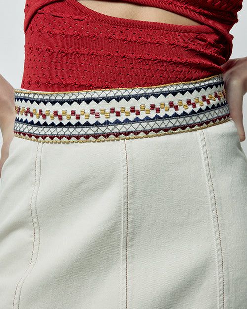 A close-up waist level view of a model wearing a red mesh top tucked into a white midi skirt with an embroidered waistline. The waistline is made up of square and triangular geometrical designs in blue, red and yellow colors with long seams.