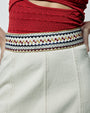 A close-up waist level view of a model wearing a red mesh top tucked into a white midi skirt with an embroidered waistline. The waistline is made up of square and triangular geometrical designs in blue, red and yellow colors with long seams.