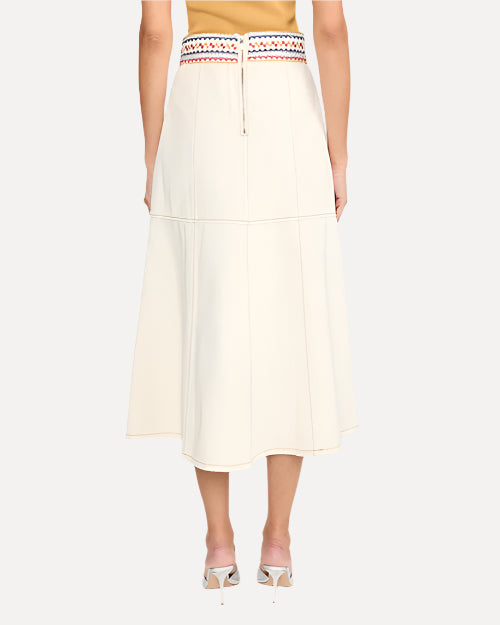 A waist down backside view of a model wearing a mustard yellow top tucked into a white midi skirt with an embroidered waistline including triangular and square geometric designs in blue, yellow and red colors. The skirt has a zipper on the back ending at the mid-flank, and the model is wearing silver open-toed heels to match.