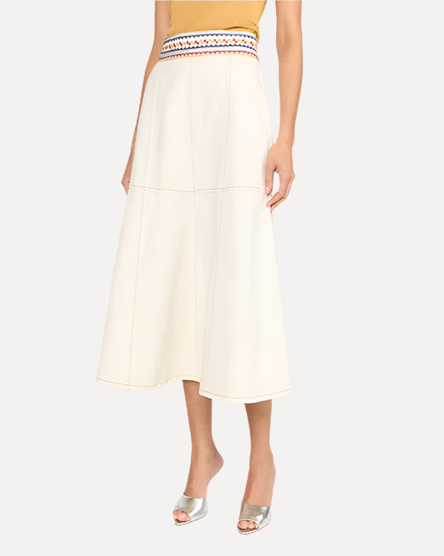 A waist down image of a model wearing a mustard yellow top tucked into a white midi skirt with an embroidered blue and red waistline. The beltline is embroidered with triangular and square geometric designs, and the model is wearing silver open-toed heels to match.